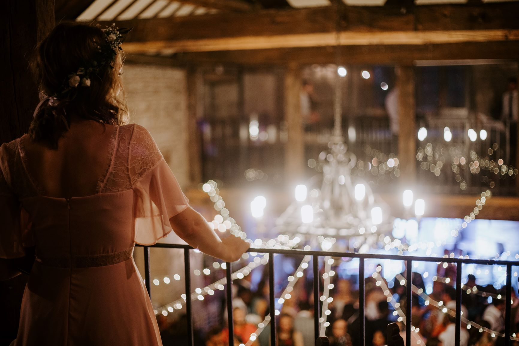 A bride is standing on a balcony looking away from the camera over a room laid out for a reception